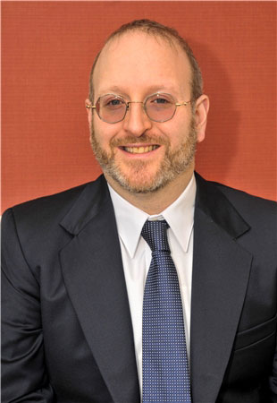 The image shows a man with glasses, smiling at the camera. He has short hair and is wearing a suit and tie.