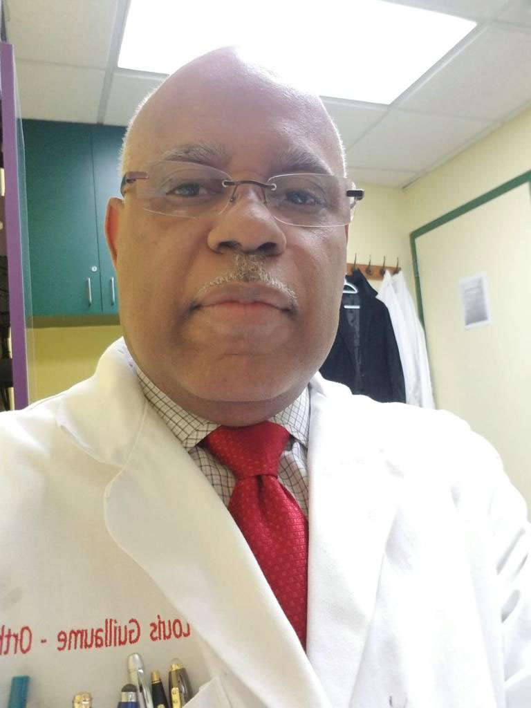 The image shows a man wearing glasses, a red tie, and a white lab coat standing in an indoor setting that appears to be a professional office or laboratory.