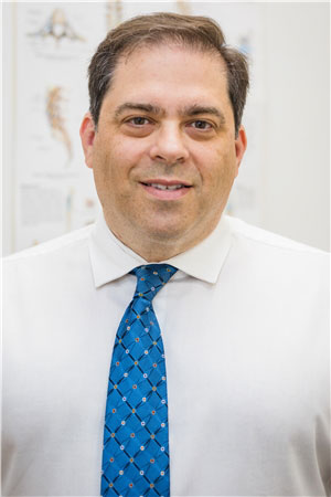 The image is a portrait of a man wearing a blue tie and standing in front of a backdrop with what appears to be medical diagrams or posters.