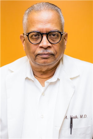 The image shows a man wearing glasses, with white hair and a mustache, dressed in a white lab coat. He is standing against a yellow background.