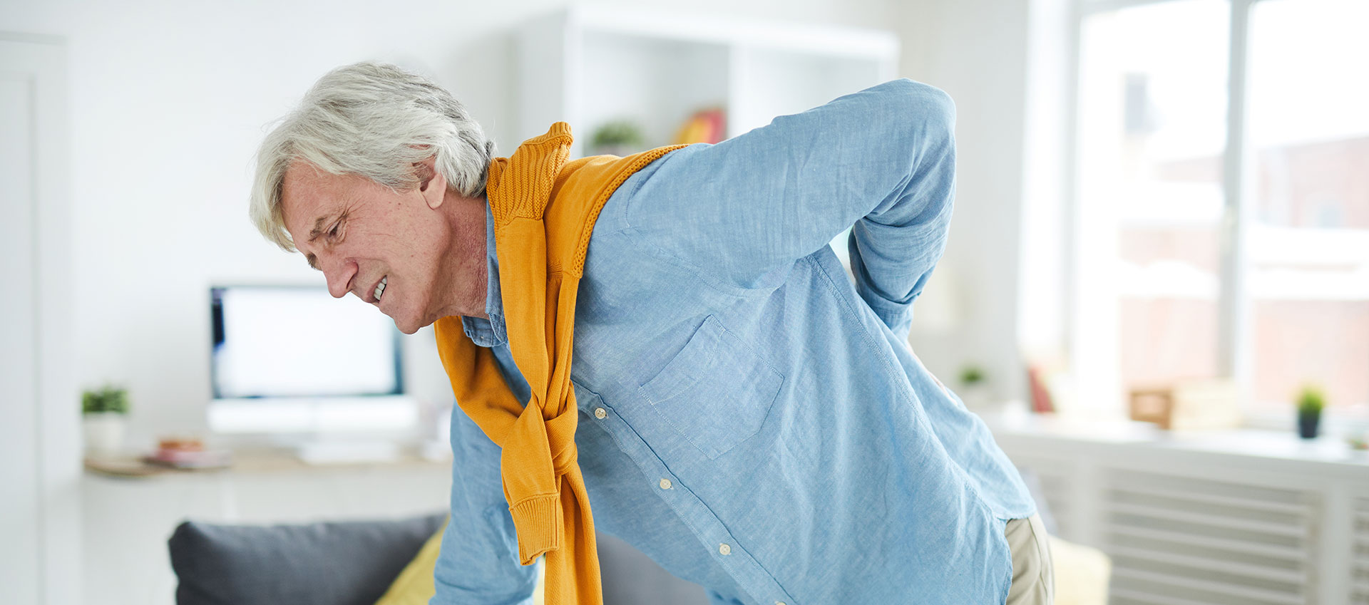 A man in a blue shirt and yellow scarf is stretching his back, standing in a living room with white walls.