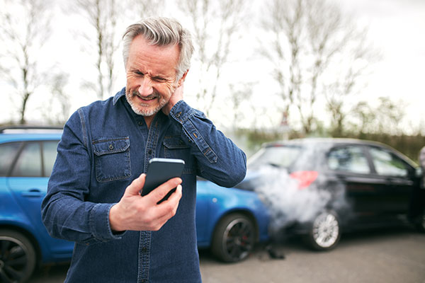 The image shows a man standing outdoors, holding a smartphone, with smoke emerging from a car in the background.