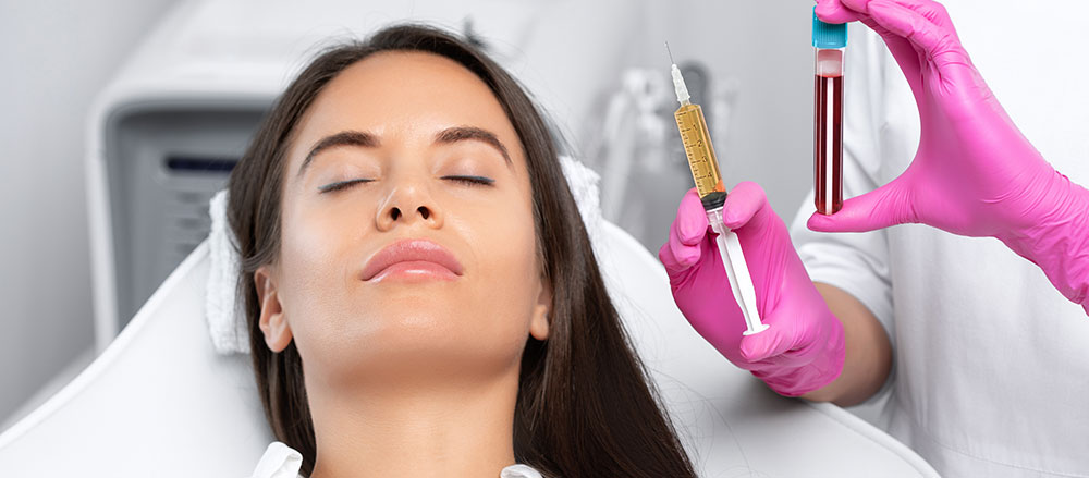 This is a photograph of a woman receiving a facial treatment, where a cosmetic professional applies a substance to her skin using a dropper, while the background suggests a medical or beauty spa setting.