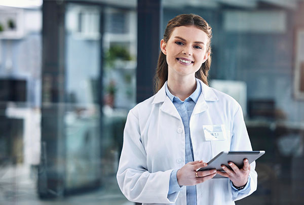 A young woman in a white lab coat, holding a tablet and smiling, stands against the backdrop of an office.