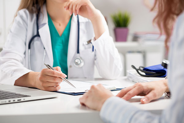 A doctor sitting at a desk, writing on a clipboard while being attended to by a patient.