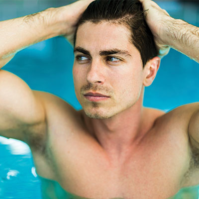 The image shows a young man with short hair, wearing a swimsuit, sitting in a swimming pool with his head resting on one hand.