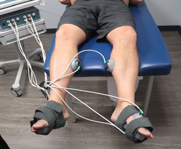 An adult male lying on a medical examination table with his feet strapped into stirrups, receiving an electrical treatment to his lower legs.
