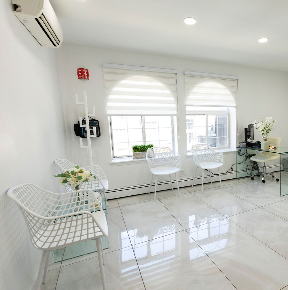 The image shows an interior space that appears to be a modern, well-lit office or waiting room. It features a clean and minimalist design with white walls, tiled flooring, and a large window allowing natural light into the room. There are two chairs with a glass table in between them, suggesting a seating area for visitors or clients. The space is furnished with a few decorative elements such as a potted plant, a small desk with a chair, and some framed artwork on the wall. The overall aesthetic is contemporary and professional.