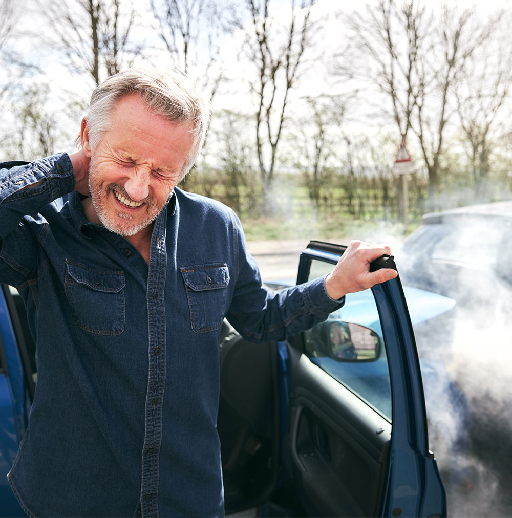 The image shows a man standing in the back of a car with a relaxed posture, wearing a blue denim shirt and jeans, smiling slightly while looking over his shoulder.