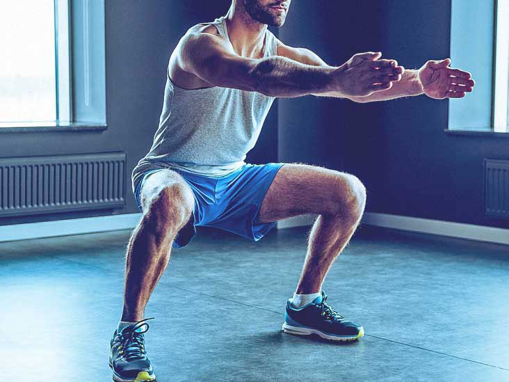 A man in a gym, performing a squat exercise with his hands on the floor, wearing athletic attire and sneakers.